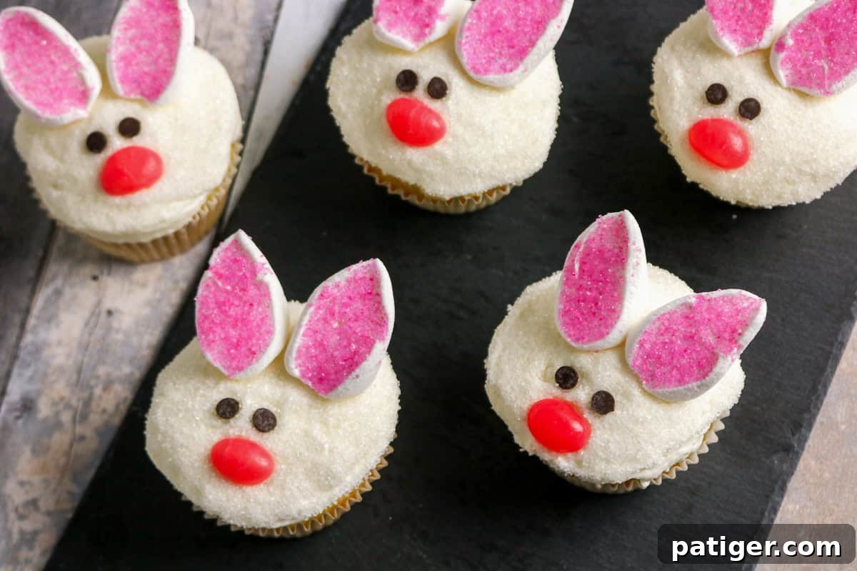 Fluffy Marshmallow Bunny Cupcakes 3 Five Easter bunny cupcakes decorated as bunny faces. Each cupcake has white sugar-coated frosting, marshmallow ears with pink sugar centers, mini chocolate chip eyes, and a red jelly bean nose, arranged on a rustic wooden board.