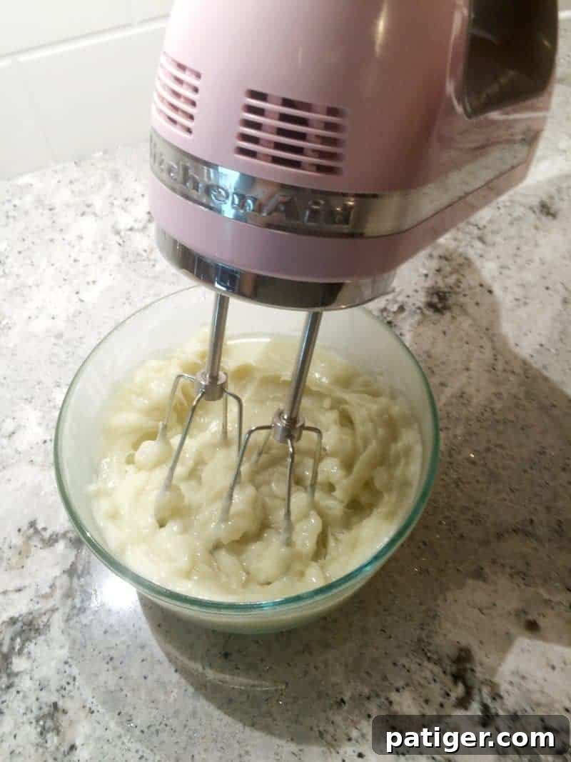 Electric hand mixer actively whipping the partially solidified body butter mixture in a glass bowl, illustrating the transformation into a light and fluffy consistency.