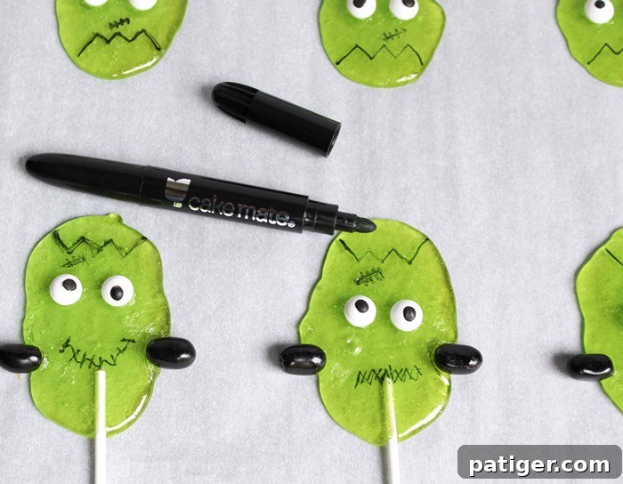 Using an edible marker to draw hair, a mouth, and a scar on the Frankenstein lollipops, completing their look.