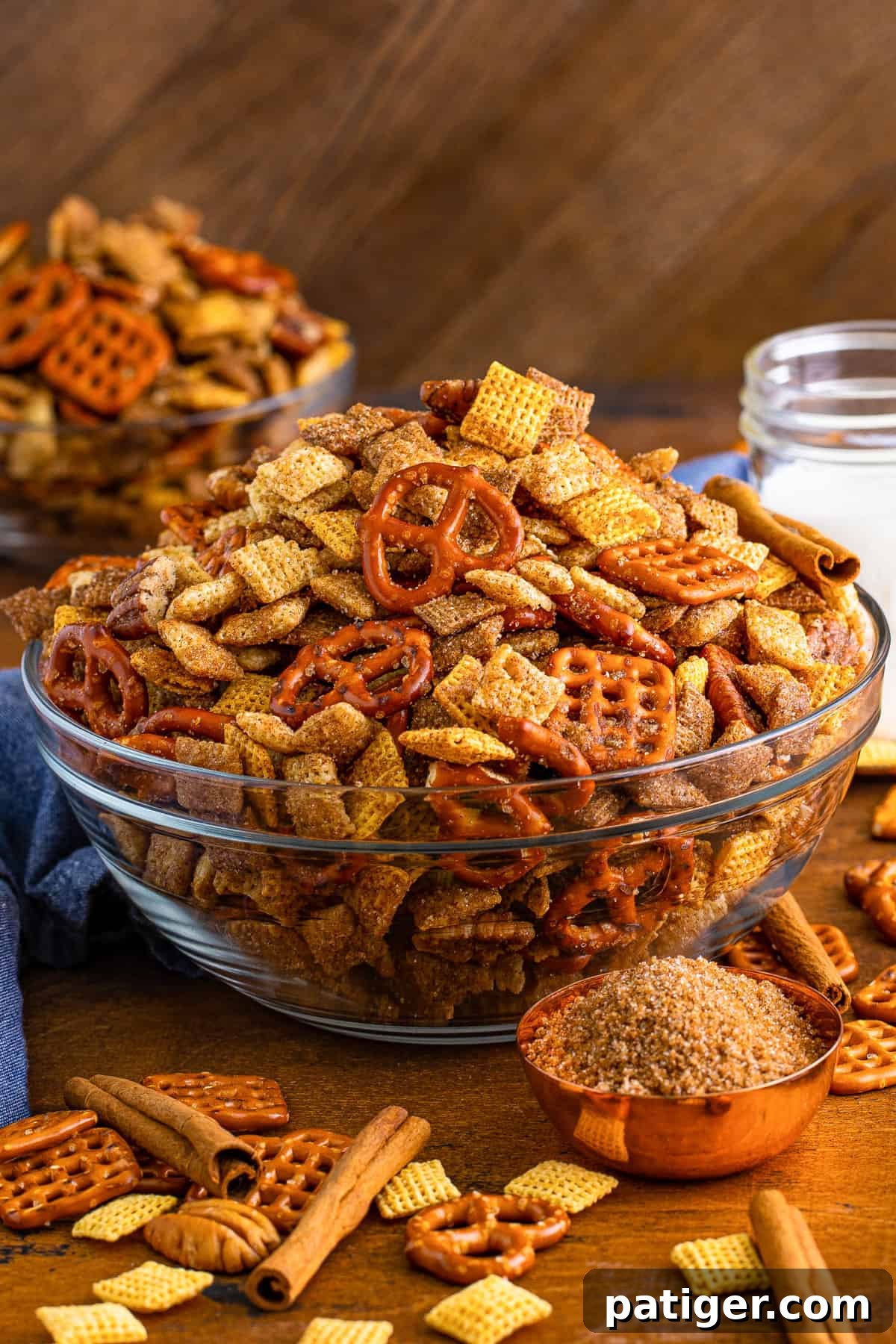 Cinnamon sugar chex mix in glass serving bowl with measuring cup of brown sugar and cinnamon sticks beside it.