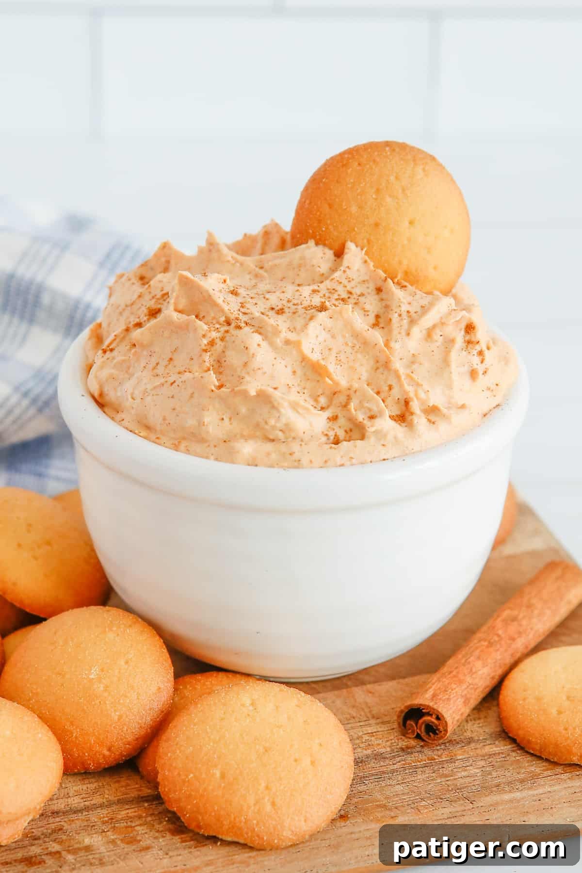 Pumpkin dip in white bowl with vanilla wafer sticking out of the top. Vanilla cookies and cinnamon sticks are on tabletop around the bowl.