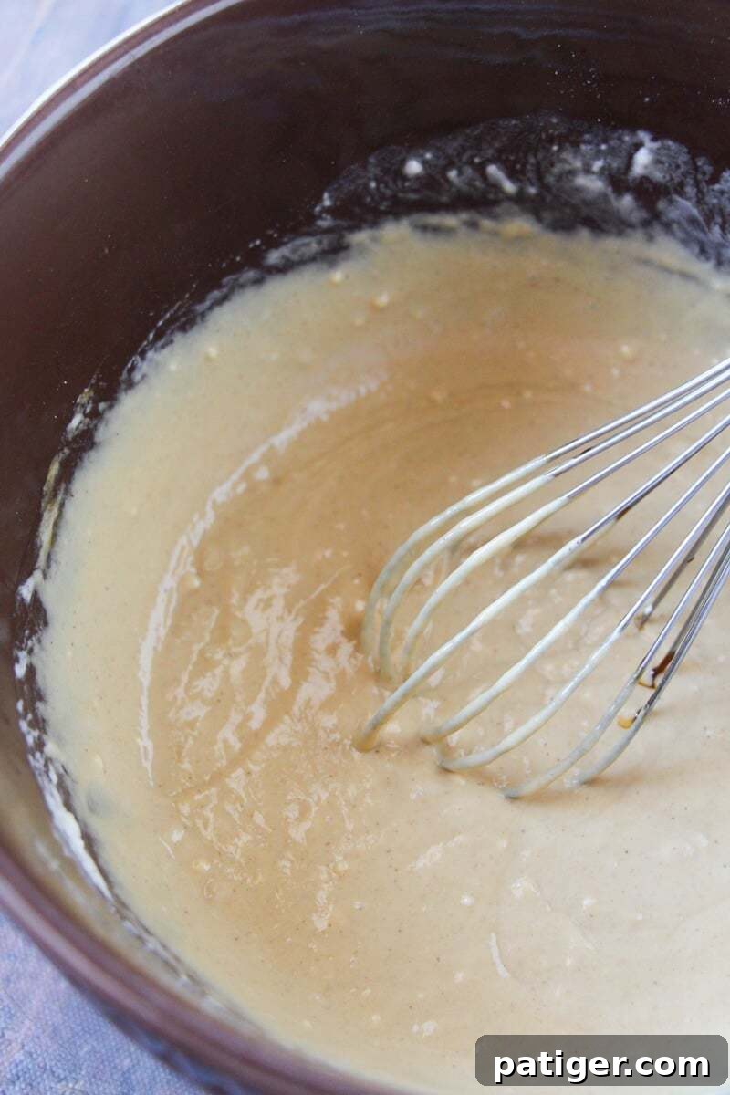 A whisk vigorously mixing wet ingredients like eggs, milk, molasses, and melted butter in a separate bowl to form the liquid base for gingerbread pancakes.