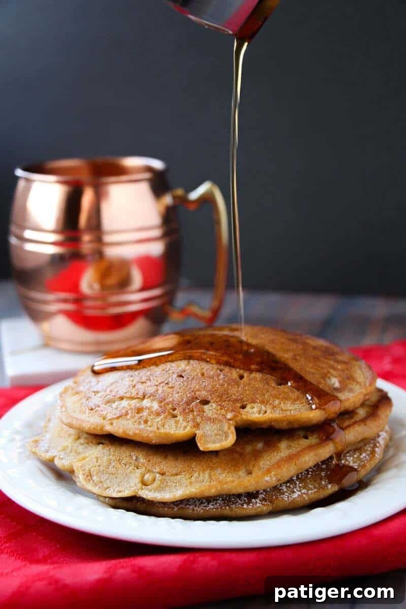 Maple syrup generously drizzled over a stack of warm gingerbread pancakes on a white plate, with a rustic bronze mug in the background.