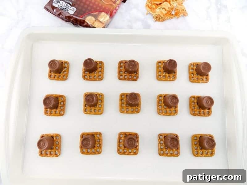 A close-up view of square pretzels on a baking sheet, each topped with an unwrapped Rolo candy positioned on one corner.