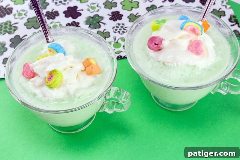 Shamrock Swirl Frozen Hot Chocolate 3 Close-up of St. Patrick's Day Shamrock Frozen White Hot Chocolate in a clear mug, garnished generously with cool whip and a variety of colorful Lucky Charms marshmallows, set against a festive green backdrop.