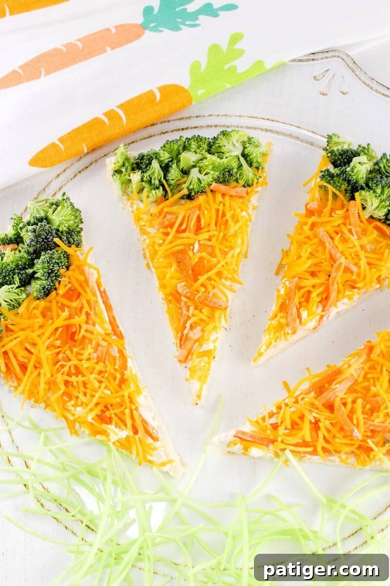Close-up of carrot-shaped veggie pizza bars, featuring a golden crescent roll base, a creamy white topping, vibrant shredded carrots, and green broccoli florets. They are arranged on a white serving dish.