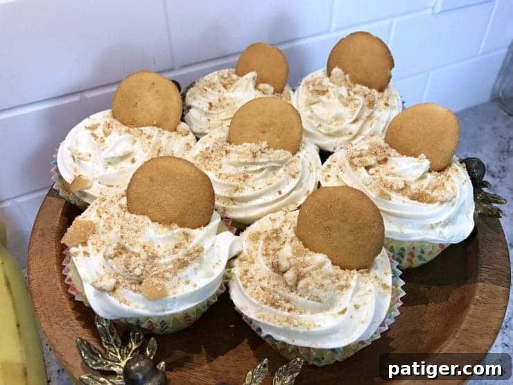 Vanilla Wafer Banana Pudding Cupcakes 2 Beautifully arranged banana pudding cupcakes, each topped with a whole Nilla wafer, presented on a rustic wooden cake stand against a soft, inviting background.
