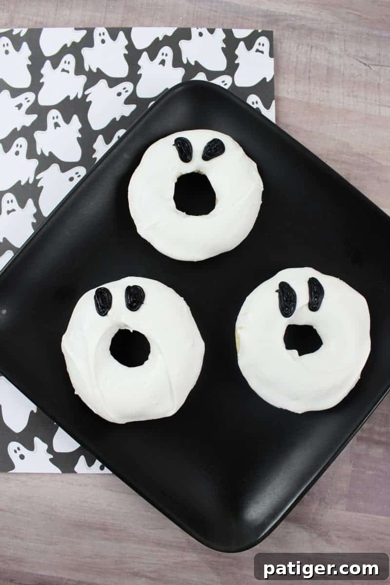 Close-up of three ghost doughnuts on a white plate, showcasing the simple black oval eyes on white frosting. Easy Halloween treat idea.