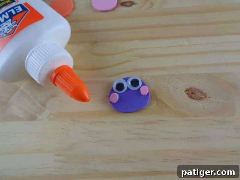 The caterpillar's head is being assembled, with two pink craft foam circles glued as cheeks and googly eyes added for expression.