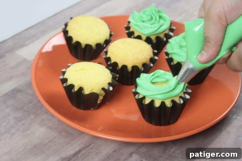 Green frosting being piped onto a vanilla cupcake using a piping bag with a large star tip.