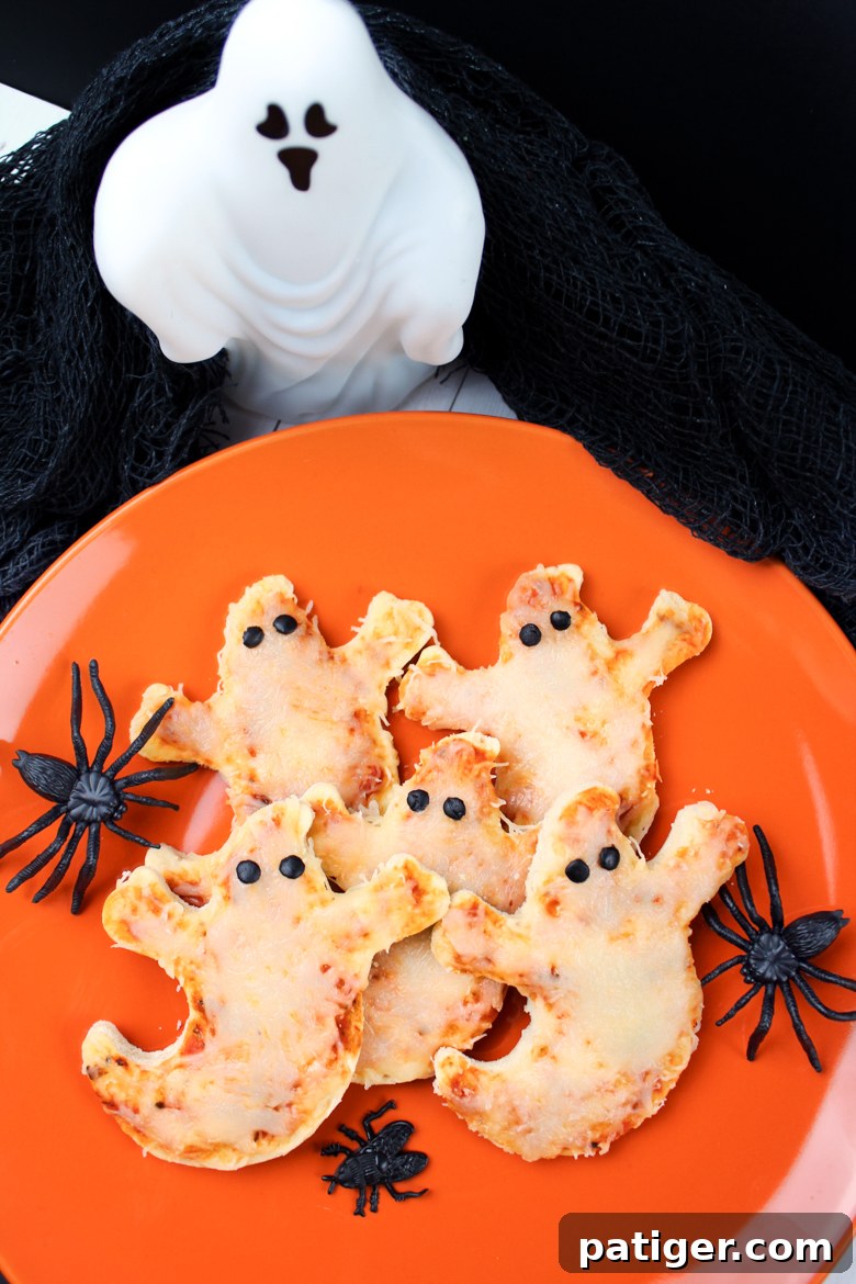 Phantom Pizza Bites 2 Ghost shaped pizzas with olive eyes served on an orange plate surrounded by Halloween decorations.