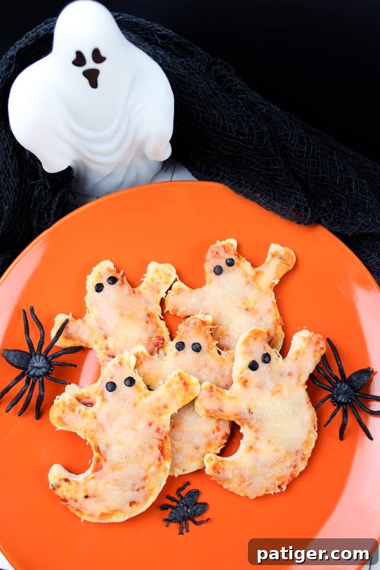 Phantom Pizza Bites 9 Ghost shaped pizzas with olive eyes served on an orange plate surrounded by Halloween decorations.