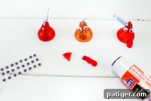 Three Hershey’s Kisses on a white surface: one plain, one with googly eyes, and one with eyes and a red felt wattle. Nearby are small googly eyes, orange felt triangles for beaks, red wattles, and a glue stick.