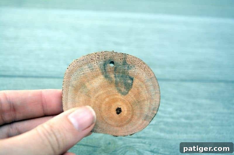 A close-up view of an adult hand drilling a small hole into the top edge of a rustic wood slice, preparing it for a ribbon hanger for a DIY ornament.