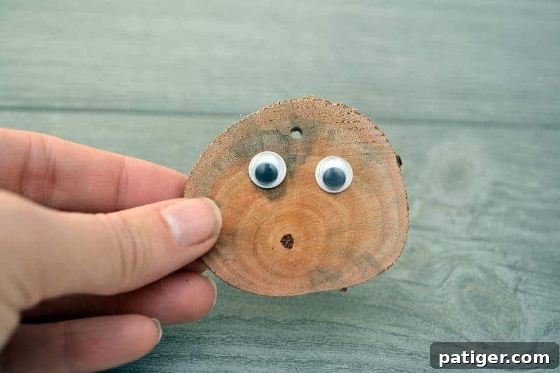 A hand applying craft glue to the back of two small googly eyes, preparing to attach them to the front of a rustic wood slice, creating the reindeer's face.