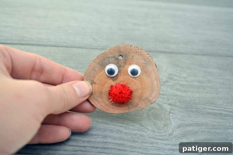 A hand pressing a sparkly red craft pom-pom onto the front of a wood slice ornament, directly below the two googly eyes, forming Rudolph's famous red nose.