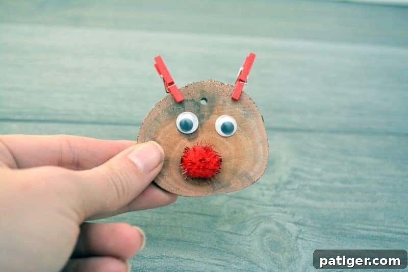 Two miniature red clothespins are attached to the top edge of a wood slice ornament, one above each googly eye, serving as the reindeer's cute antlers.