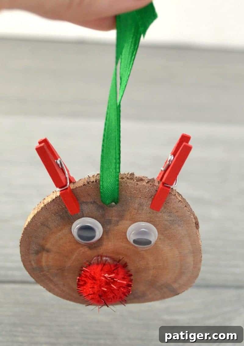 A close-up of a hand threading a piece of red ribbon through a pre-drilled hole at the top of a finished wood slice Rudolph ornament, creating a loop for hanging.