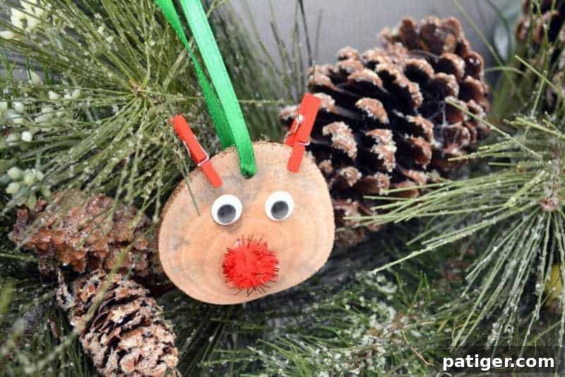 A completed DIY rustic wood slice Rudolph the Red-Nosed Reindeer ornament, showcasing its googly eyes, sparkly red nose, and red clothespin antlers, ready to be hung on a Christmas tree.
