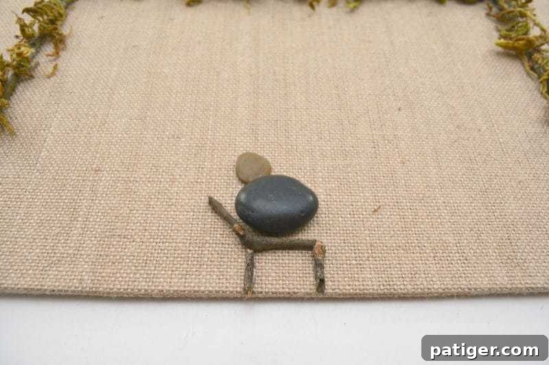 Pebble Art Nativity Canvas 6 Create Baby Jesus using a medium flat stone for the body and a small pebble for the head, then glue him into place with a twig manger below on the canvas.