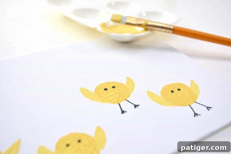 A paintbrush applying a small, curved yellow wing shape to the side of a cork-stamped chick body on a canvas.