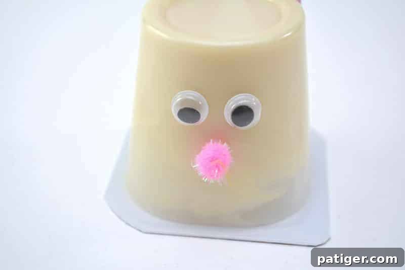 The action of attaching a small pink pom pom to serve as the bunny's nose on the Easter Bunny Pudding Cup, adding a soft, textured and adorable detail.