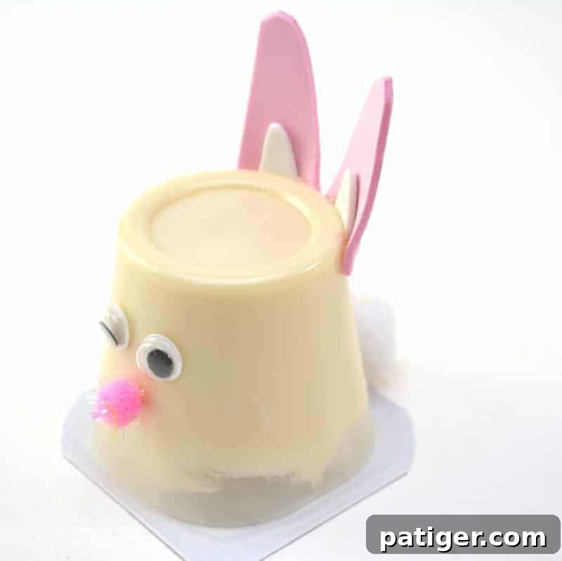 Gluing a medium white pom pom to the backside of the pudding cup, carefully creating a fluffy and charming tail for the Easter Bunny craft.