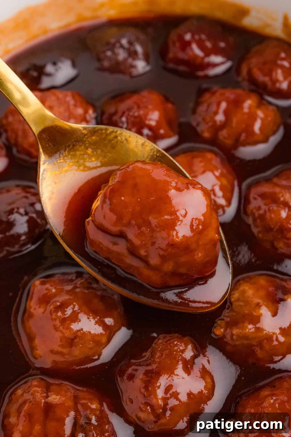 A gold spoon holds a barbecue and grape jelly-coated meatball, lifted from the slow cooker. The thick sauce drips slightly, showing its rich consistency. Other meatballs remain submerged in the sauce in the background.
