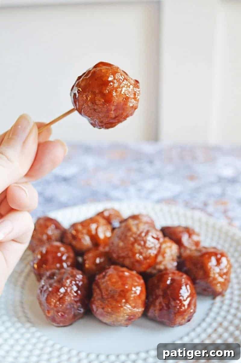 Meatballs with grape jelly and bbq sauce on a plate with a single meatball picked up with a hand holding a tooth pick.
