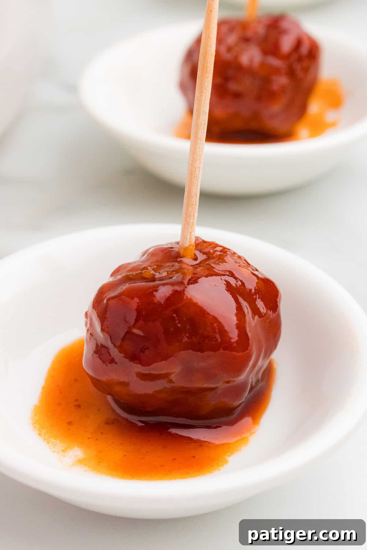 A crockpot meatball covered in thick barbecue and grape jelly sauce sits in a small white dish. A wooden toothpick is inserted in the centerA crockpot meatball covered in thick barbecue and grape jelly sauce sits in a small white dish with a wooden toothpick in the center.