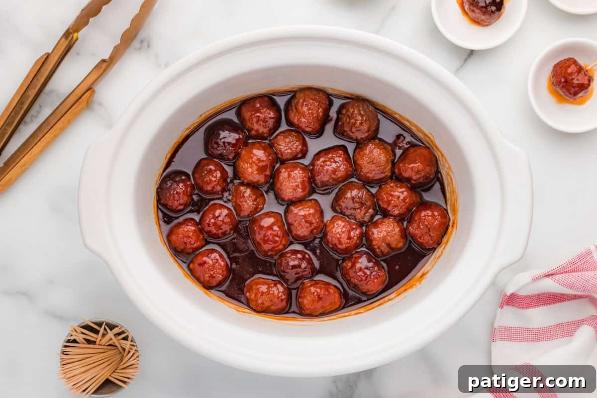 A white slow cooker filled with barbecue and grape jelly meatballs, viewed from above. The glossy sauce coats each meatball evenly. The scene includes serving plates and gold tongs on a marble countertop.