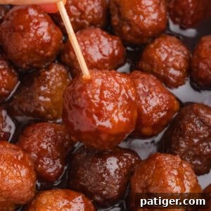 A close-up of a glazed meatball covered in thick barbecue and grape jelly sauce, held by a wooden toothpick. More crockpot meatballs are visible in the background, coated in the glossy sauce.