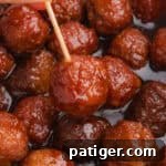 A close-up of a glazed meatball covered in thick barbecue and grape jelly sauce, held by a wooden toothpick. More crockpot meatballs are visible in the background, coated in the glossy sauce.