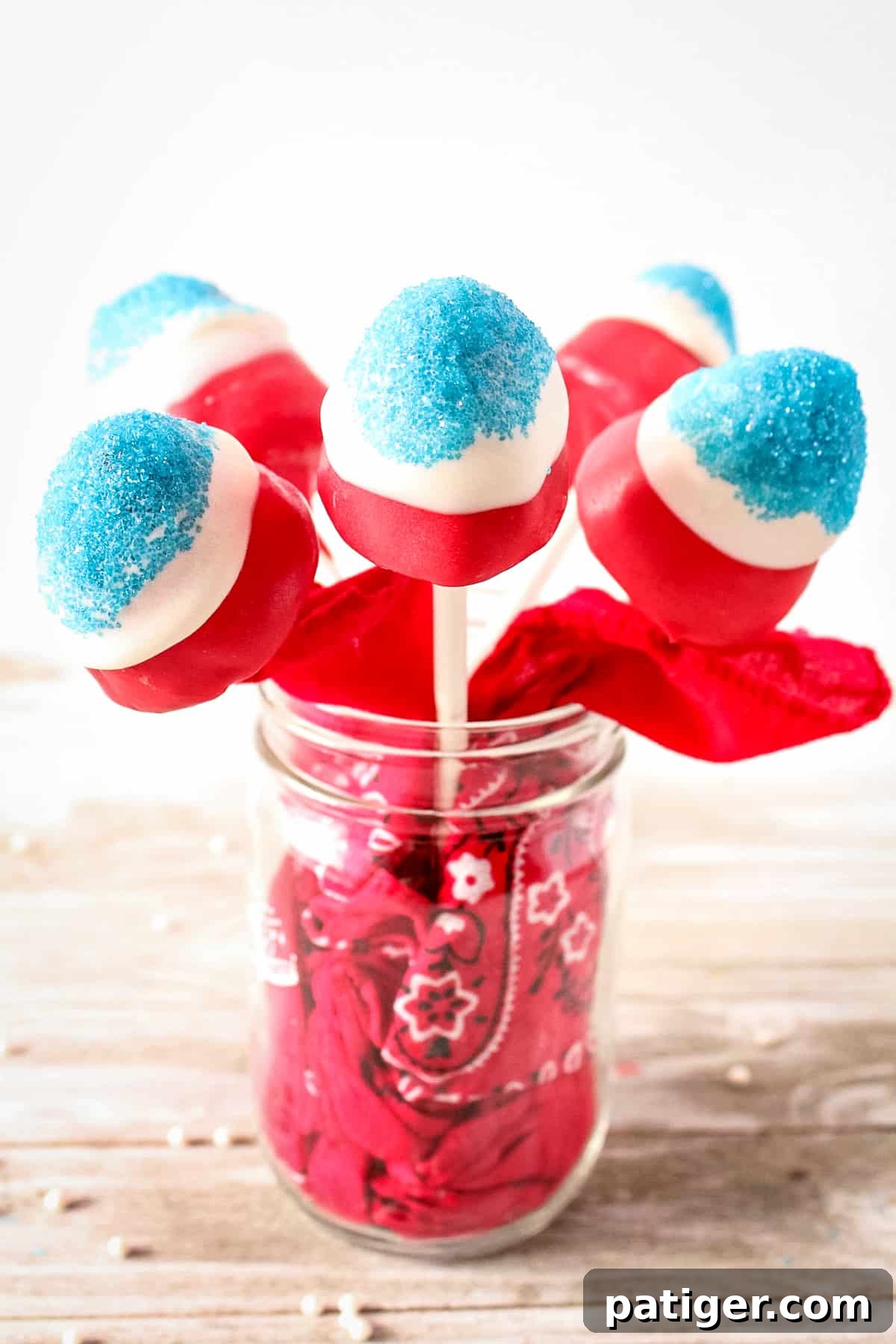 Red, white, and blue covered strawberries on sticks in a mason jar. The mason jar has a red bandana stuffed in it.