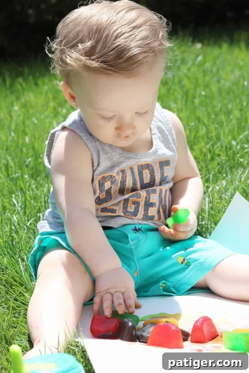 A baby's hand reaches for brightly colored ice cubes on a white poster board, showcasing a fun and stimulating sensory play activity for summer outdoor enjoyment.