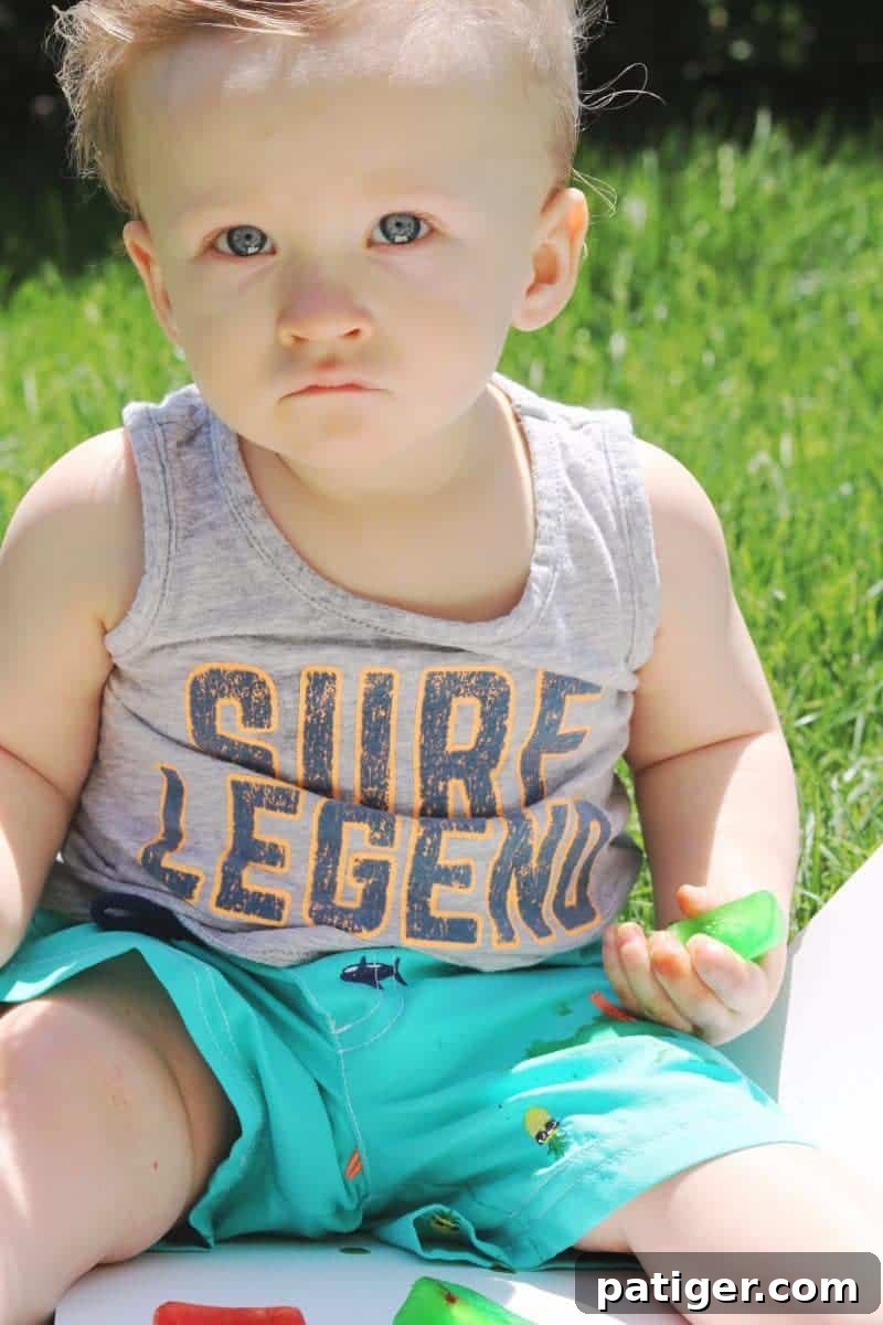 A toddler happily engages in colored ice play outdoors, exploring the vibrant cubes on a grassy lawn – a perfect sensory activity for hot summer days.