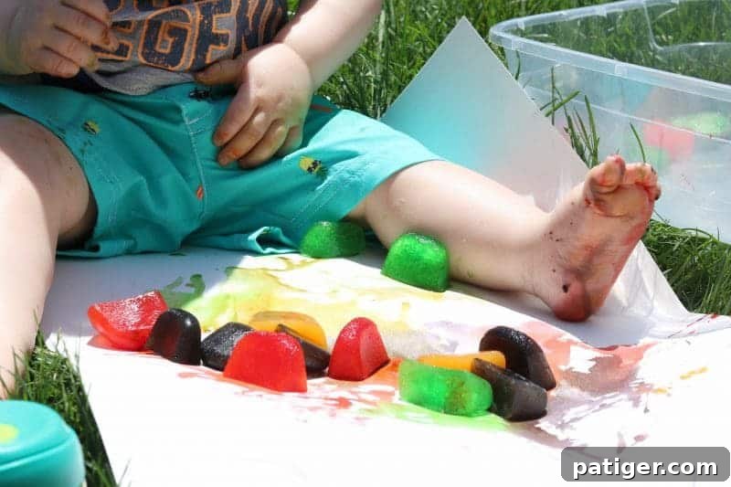 A baby's small hands explore and interact with vibrant colored ice cubes on a poster board, providing a captivating sensory experience during outdoor playtime.