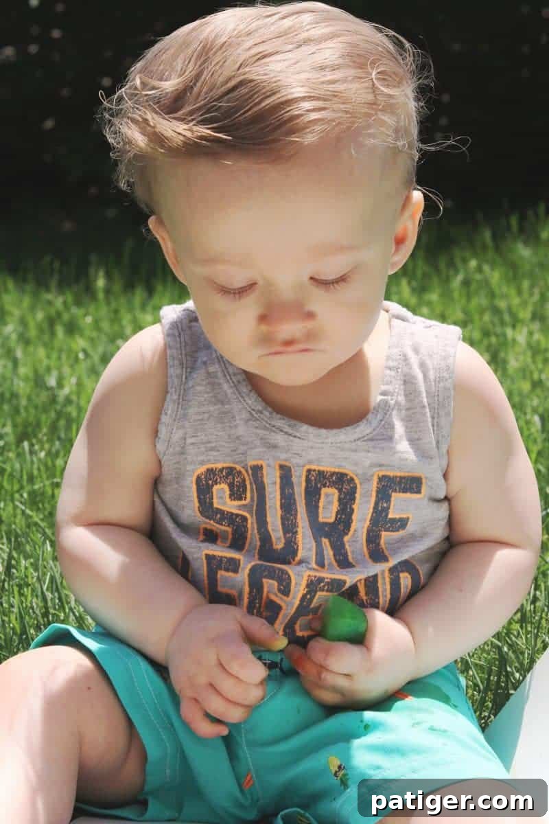 A baby's tiny hand firmly squeezes a colorful ice cube, demonstrating fine motor skill development and tactile exploration during a sensory play activity.