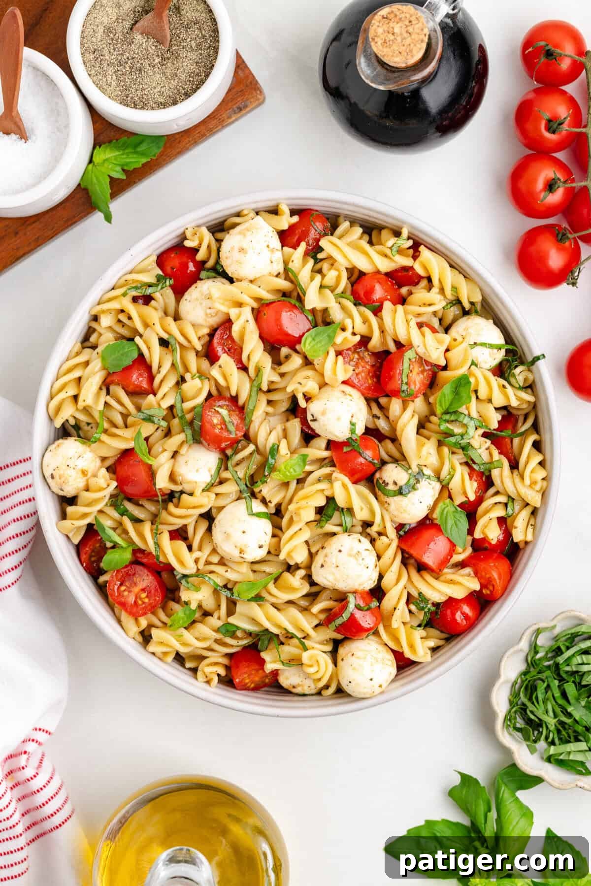 Sun-Kissed Caprese Pasta Salad 2 Overhead view of a large bowl of caprese pasta salad with rotini, cherry tomatoes, mozzarella balls, and basil. Around the bowl are a bottle of balsamic vinegar, salt, pepper, olive oil, fresh basil leaves, and cherry tomatoes on the vine.