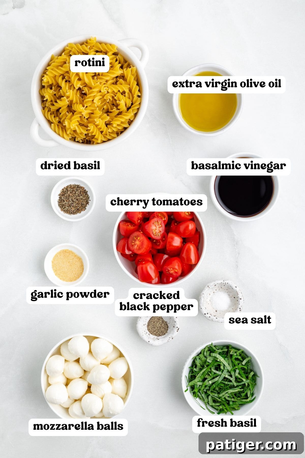Sun-Kissed Caprese Pasta Salad 4 Overhead view of labeled ingredients in bowls for making Caprese pasta salad. Labels include rotini, extra virgin olive oil, balsamic vinegar, dried basil, garlic powder, cherry tomatoes, cracked black pepper, sea salt, mozzarella balls, and fresh basil.