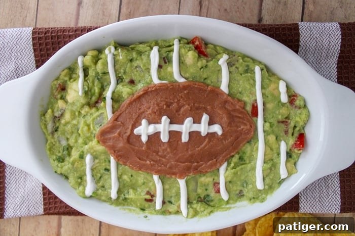 A beautifully decorated guacamole football field appetizer, complete with sour cream yard lines and a refried bean football, served with tortilla chips.