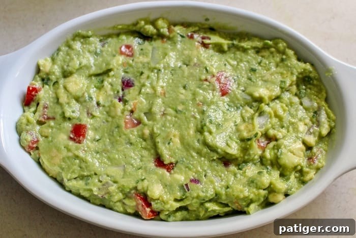 A pair of hands mixing a bowl of freshly made guacamole with a fork, showing the creamy texture and integrated ingredients. The bowl is set on a wooden surface.