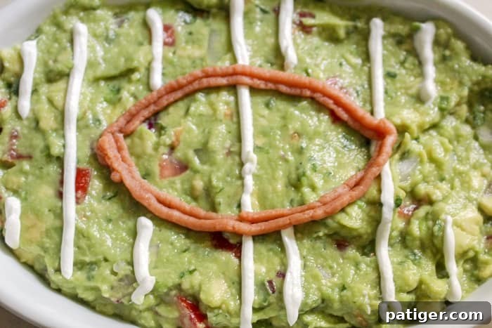 A close-up of a serving dish with green guacamole, being decorated with white sour cream piped from a plastic baggie to create the yard lines of a football field. 