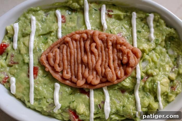 A football-shaped design made from refried beans smoothed onto green guacamole, illustrating the in-process decoration of the game day appetizer.