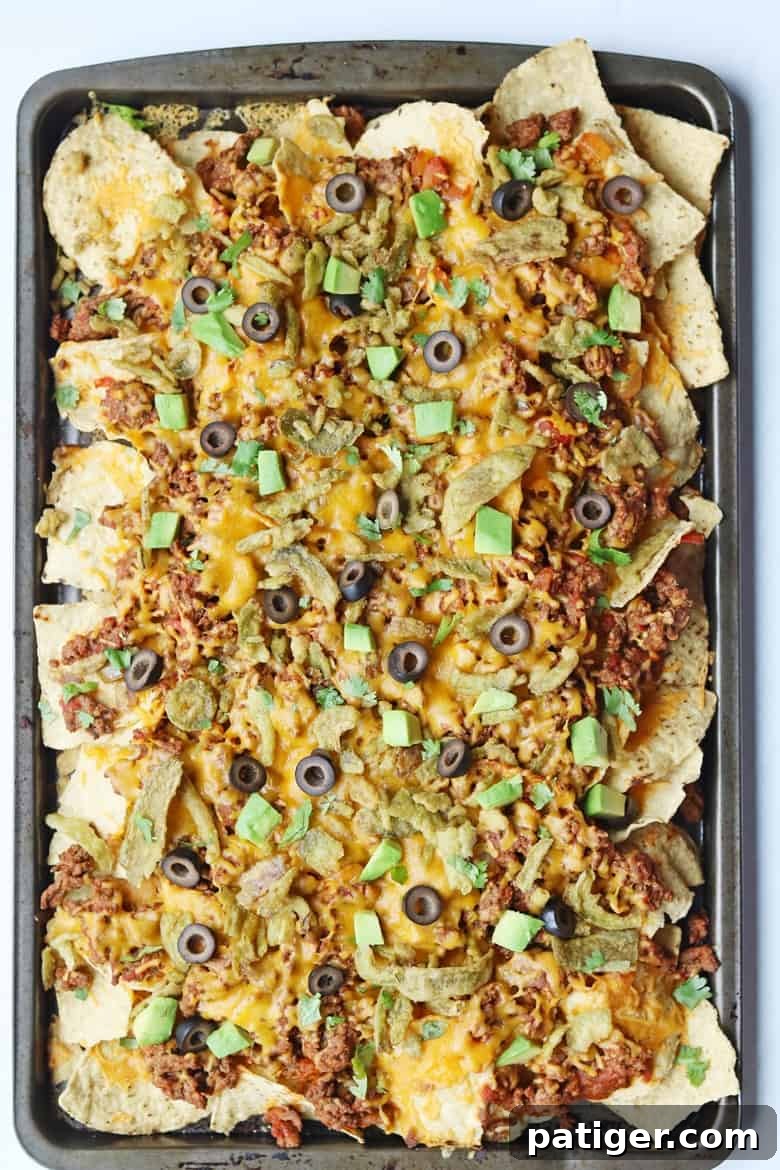 A large baking sheet of freshly baked, loaded nachos, piled high with ground beef, melted cheddar cheese, and scattered with various toppings.