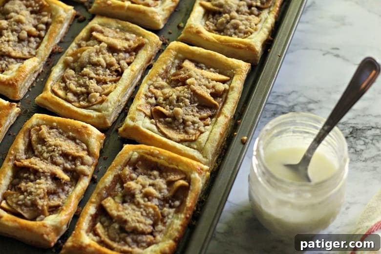 Freshly baked apple danishes cooling on a baking sheet, with a jar of icing ready to be drizzled over them.