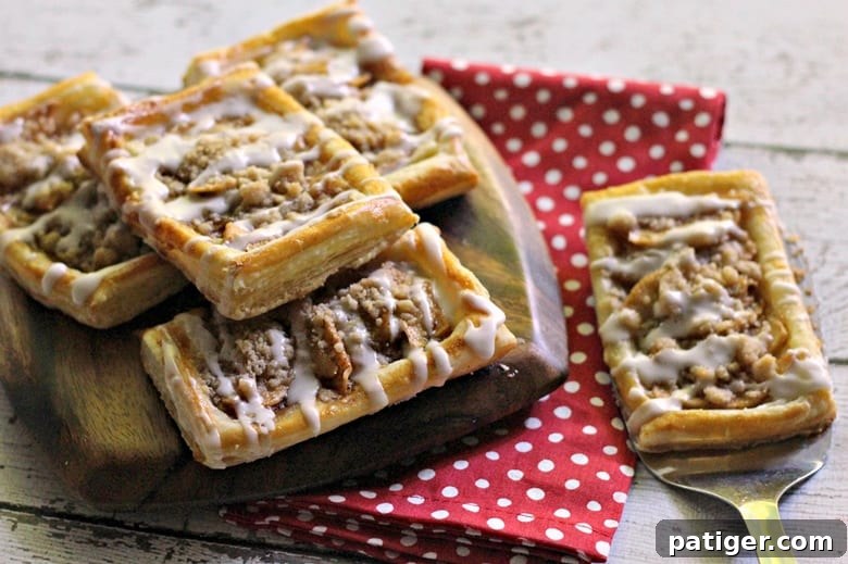 Three golden-brown apple danishes arranged on a baking sheet, showcasing their delicious crumb topping and a generous drizzle of white icing. This tasty apple crumb danish recipe offers a sweet breakfast or brunch option.