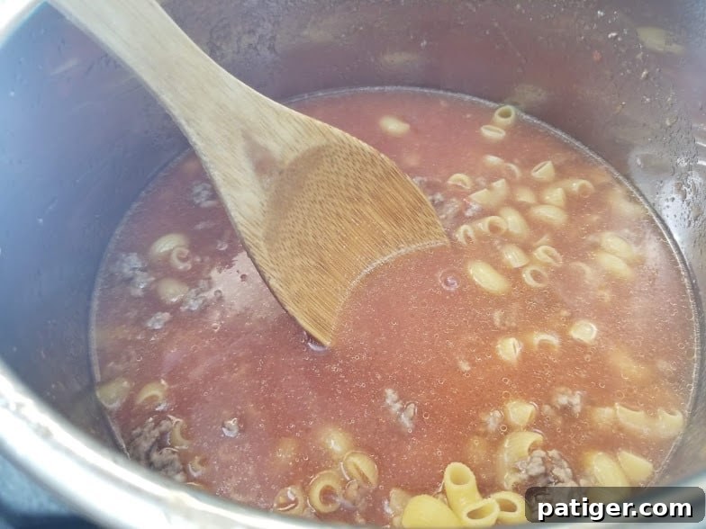 Instant Pot filled with uncooked macaroni, browned beef, beef broth, tomato puree, and seasonings, ready for pressure cooking.