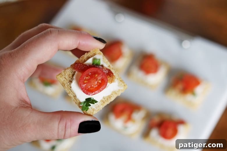 Festive Triscuit Bites 6 A single Triscuit cracker featuring a generous spread of cream cheese, topped with crispy bacon and a vibrant tomato slice.