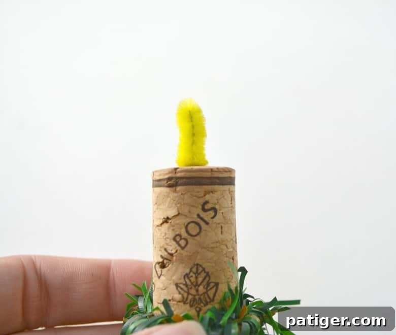 Handmade Wine Cork Candle Ornaments 6 A vibrant yellow pipe cleaner artfully shaped to resemble a flickering flame, gracefully positioned atop a wine cork already adorned with a miniature pine garland wreath.